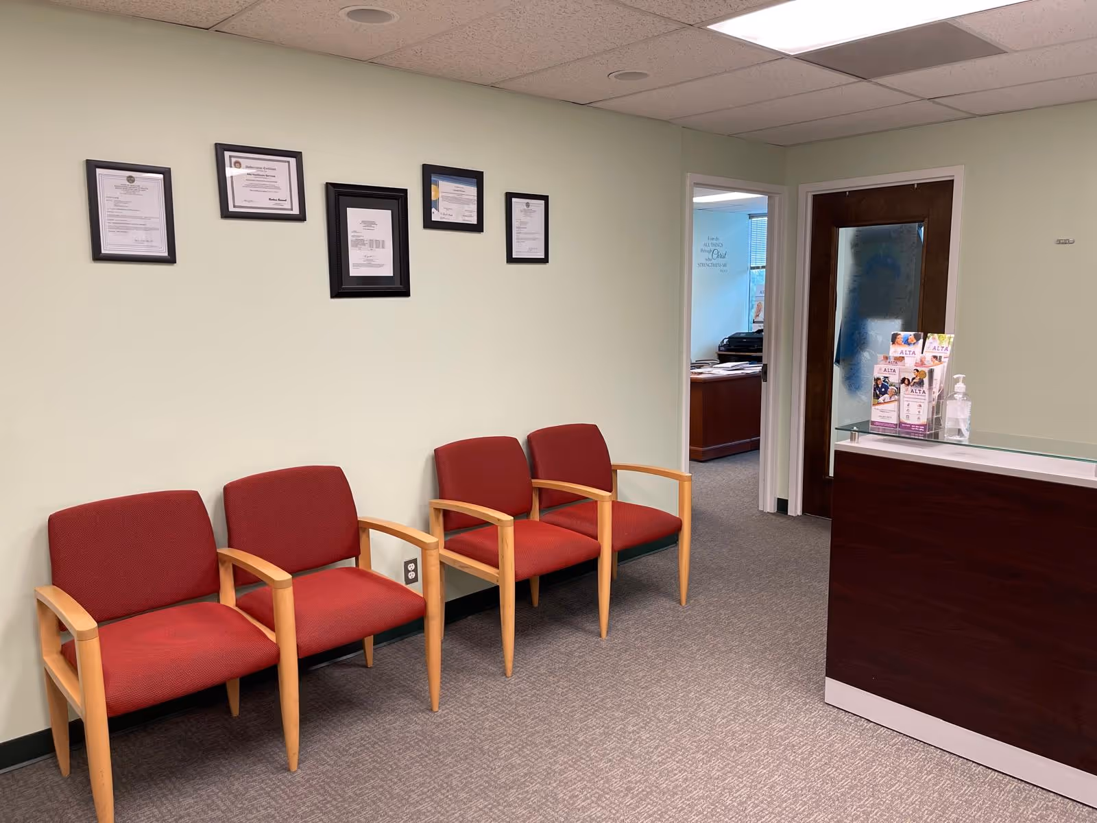 Reception area with red upholstered chairs, framed certificates on pale walls, near a dark wood desk.