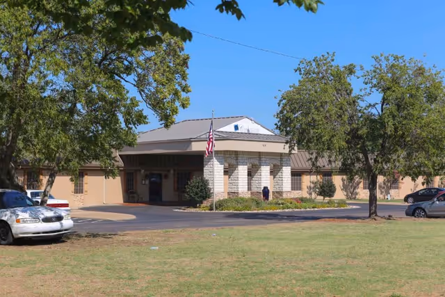 Exterior view of Carrollton Health & Rehabilitation Center building with an American flag in front, surrounded by trees and parked cars on a sunny day.