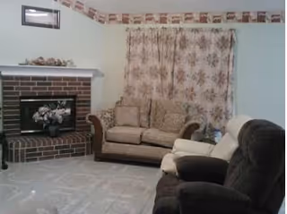 Cozy senior living facility living room with brick fireplace, floral curtains, and beige seating.