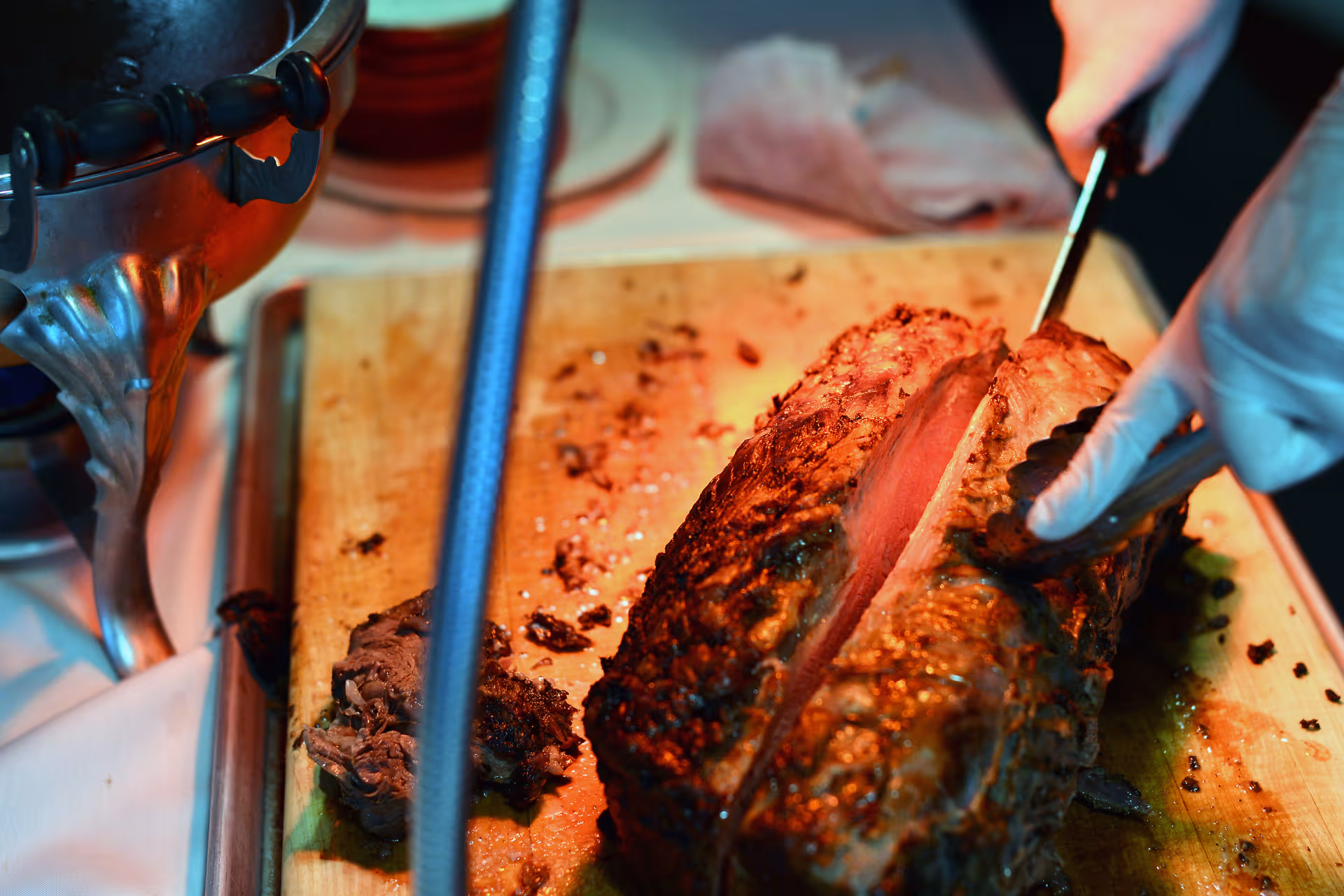 A close-up of a person wearing gloves slicing a large piece of cooked meat on a wooden cutting board with tongs and a knife.