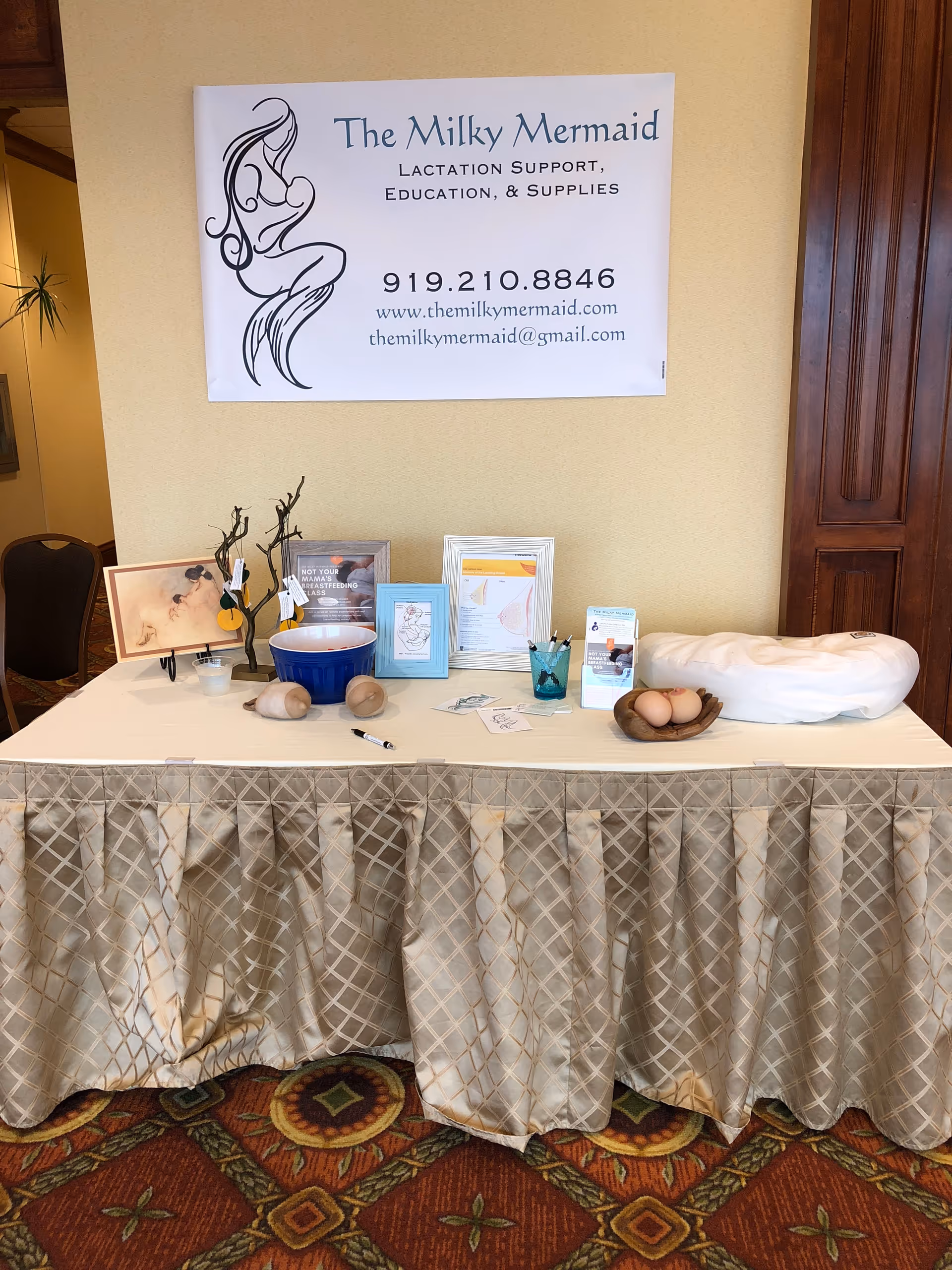 Indoor display table with promotional materials and a large The Milky Mermaid Lactation Services banner.