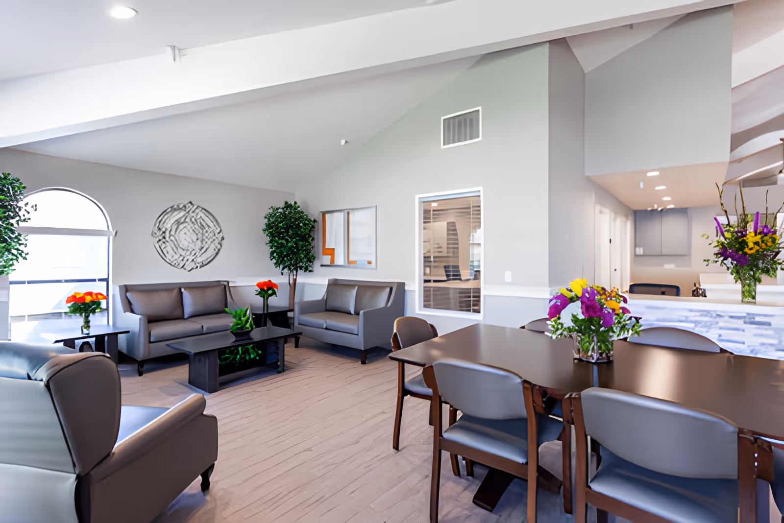 Bright communal living room and dining area with sofas, a coffee table, a large dining table, and fresh flower arrangements.