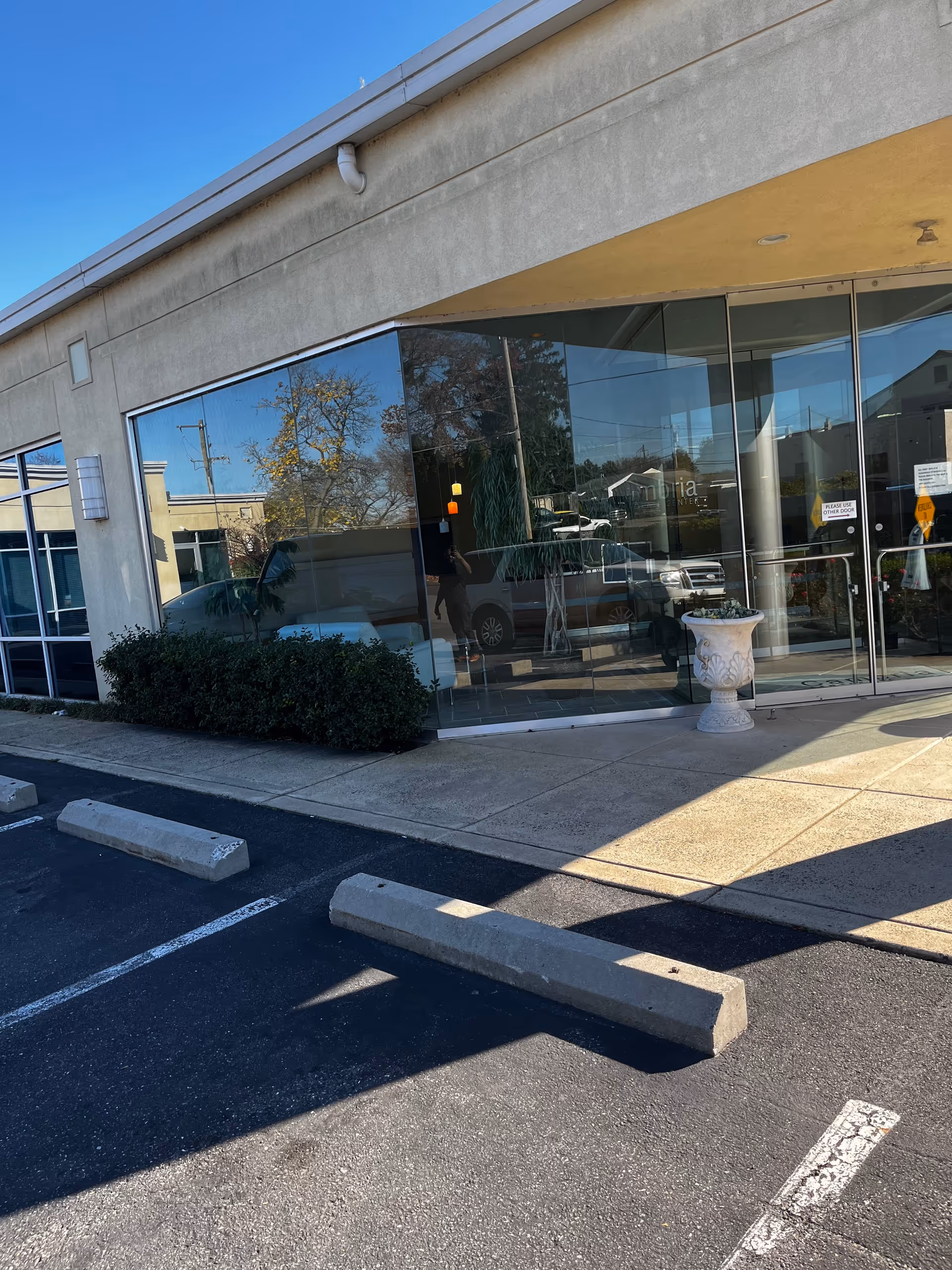 Glass storefront entrance with reflections of cars and sky, sidewalk, and adjacent parking area.