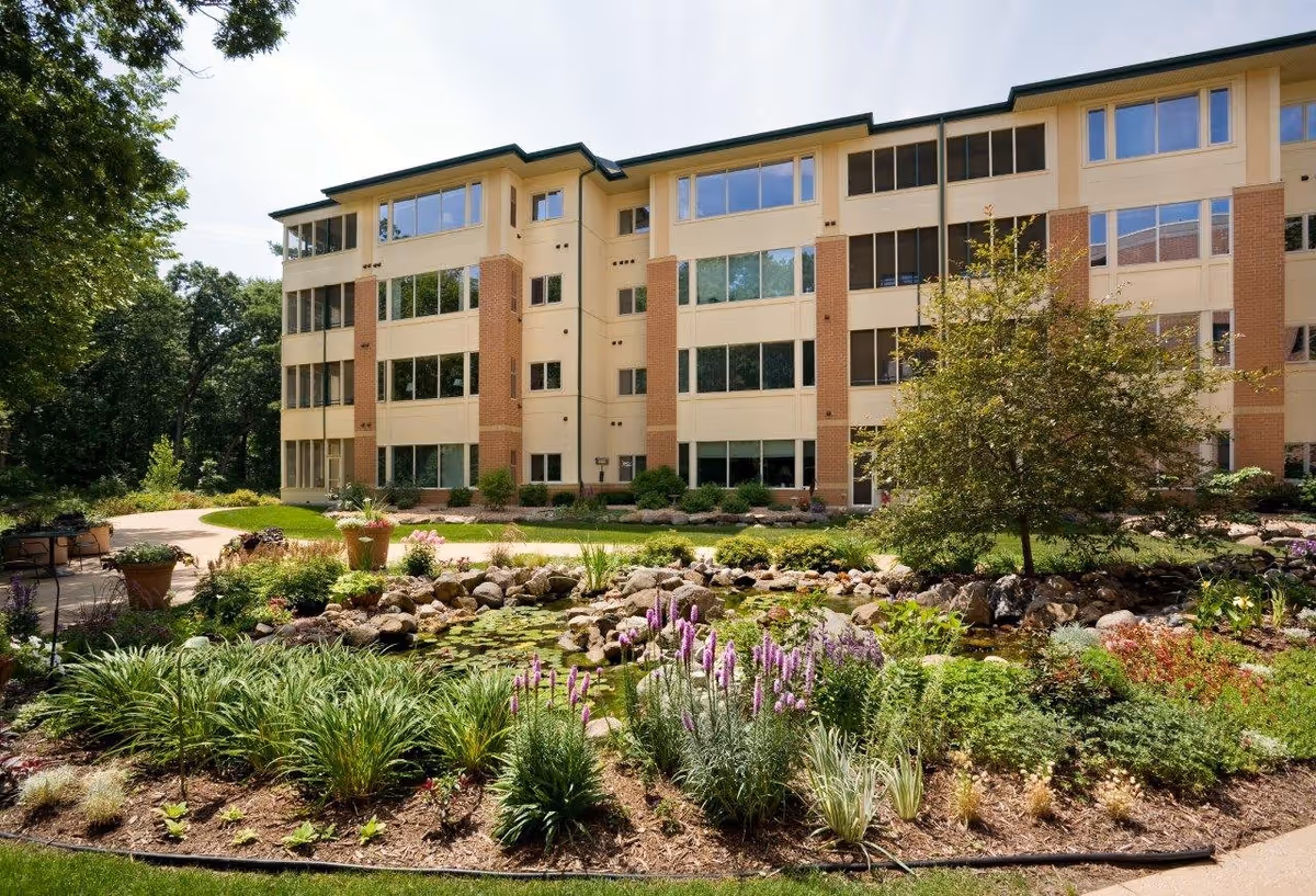 Exterior view of a multi-story senior living facility building with large windows, surrounded by a landscaped garden featuring a pond, rocks, various plants, flowers, and a small tree under a clear sky.