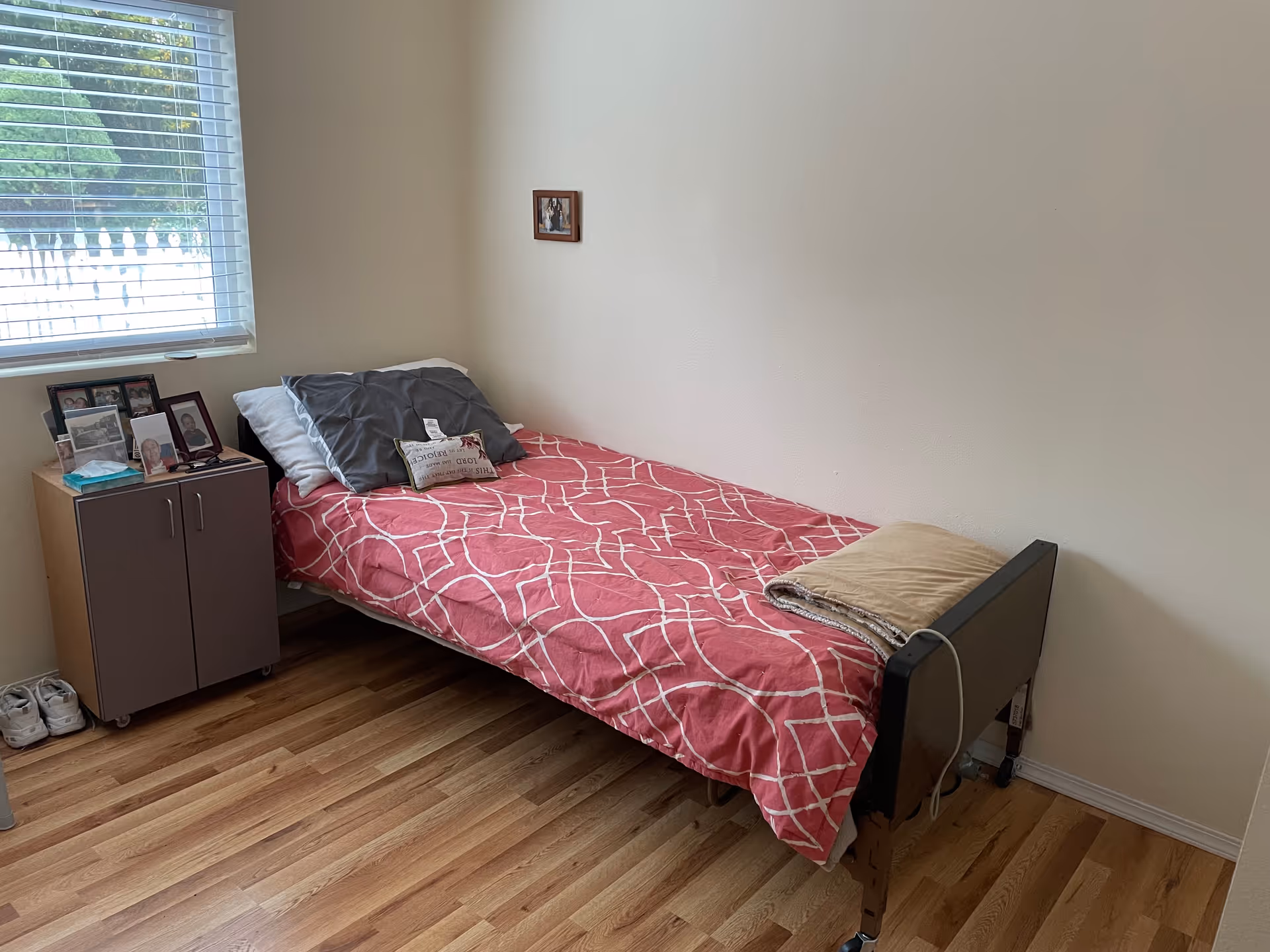 A small bedroom with a single bed, pink cover, window, and photo frames on a cabinet.