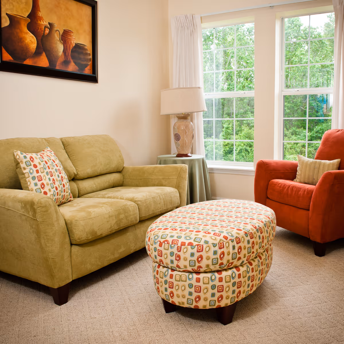 Bright living room with a green sofa, patterned ottoman, red armchair, table lamp, and large windows showing greenery.