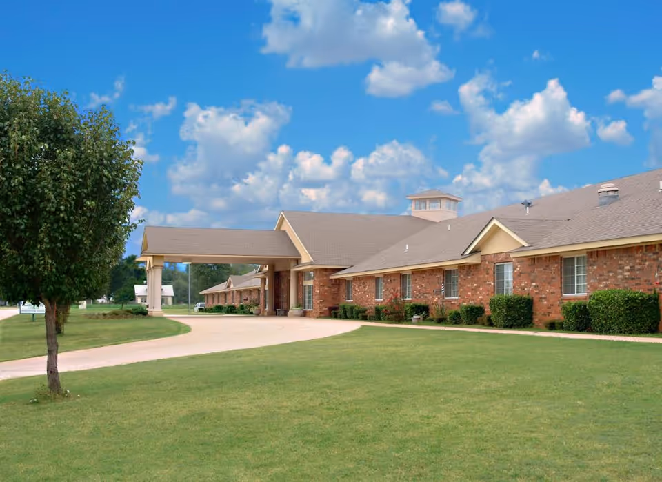 Front exterior of a single-story brick senior living building with a covered entrance, driveway, lawn, and a tree under a blue sky.
