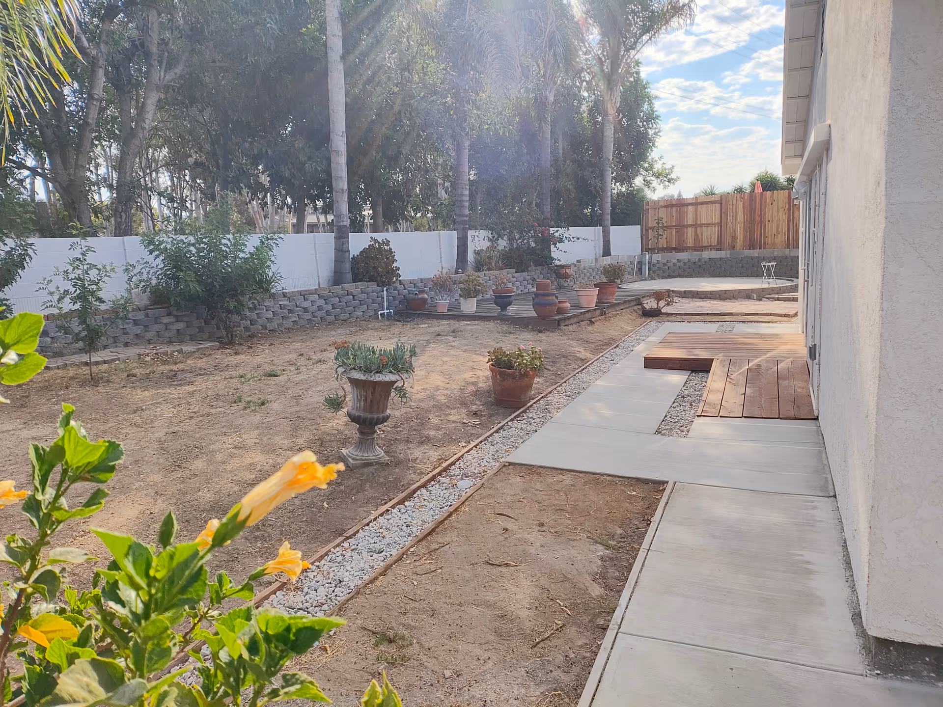 Backyard garden area with potted plants, a concrete path, and a wooden deck beside a house.
