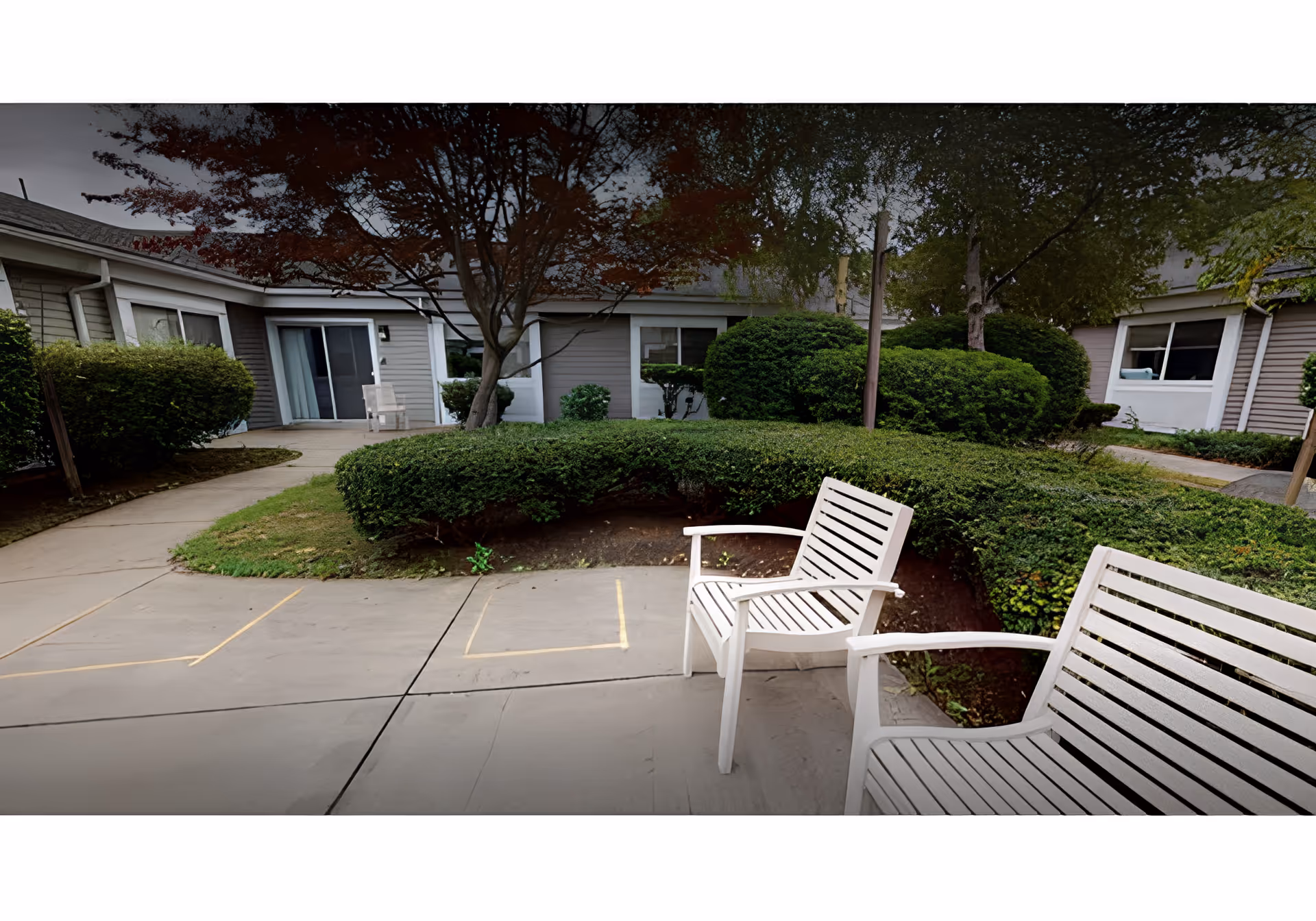 Outdoor courtyard of a single-story senior living facility with two white patio chairs, paved walkways, trimmed hedges, and building entrances.