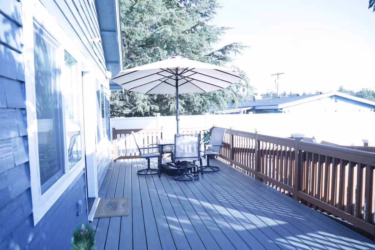 Outdoor wooden deck with a table, chairs, and a large umbrella beside a blue house.