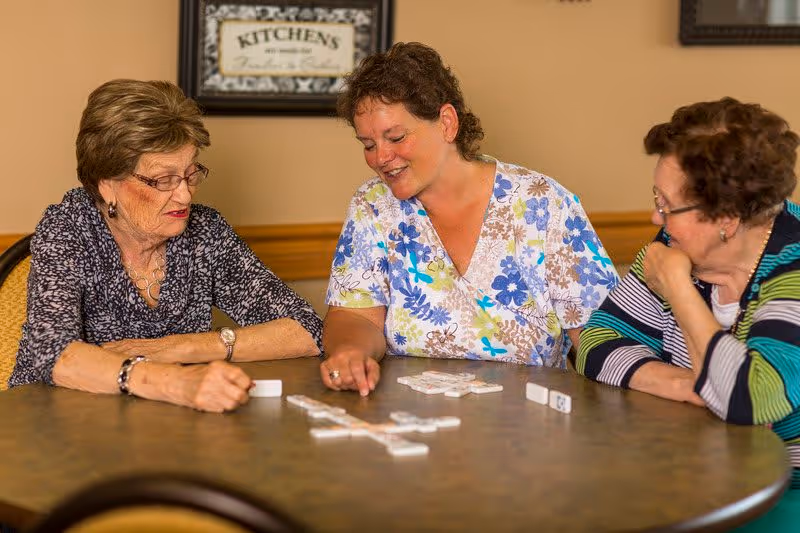 Three women sitting around a round table playing a game of dominoes in a cozy indoor setting. One woman in a floral shirt is smiling and pointing at the dominoes, while the other two women watch attentively. The background features a framed sign that reads 'KITCHEN'S are made for Friends & Food.'