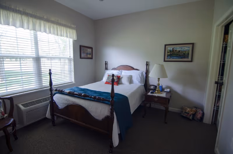 Small furnished bedroom with a wooden four-poster bed, nightstand and lamp beside a large window with blinds.