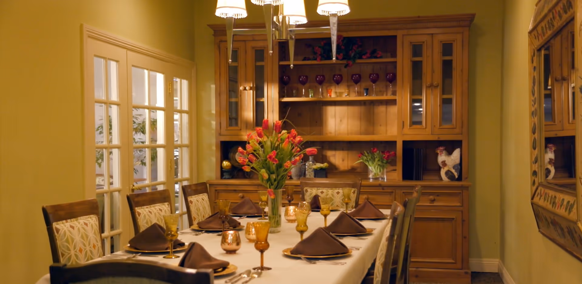 A formal dining room with a set table featuring brown napkins and glassware, a vase of red tulips as the centerpiece, and a wooden hutch in the background.