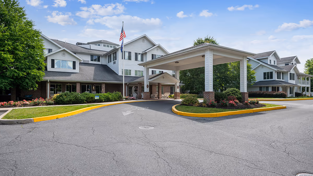 Exterior view of Holiday Essex House senior living facility showing a large driveway with a covered entrance, surrounded by landscaped greenery and flowers, with a multi-story building in the background under a partly cloudy sky.