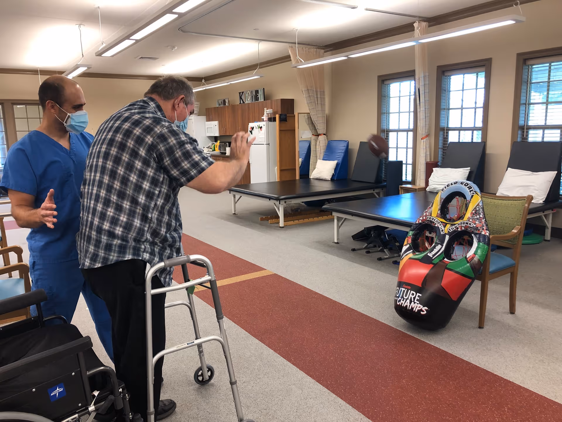An elderly man using a walker is playing a bean bag toss game with the assistance of a healthcare worker wearing blue scrubs and a face mask in a rehabilitation facility room. The room has therapy tables, chairs, and large windows with curtains.