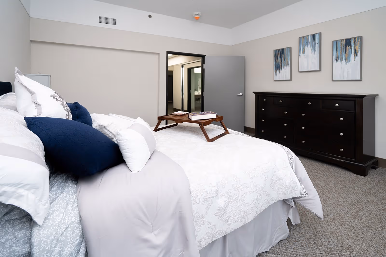 A neatly made bed with white and gray bedding and multiple pillows, including dark blue ones. A wooden tray with a book and a small item is placed on the bed. In the background, there is a dark wooden dresser with multiple drawers and three abstract paintings hanging above it. An open door reveals a glimpse of a bathroom with a sink and mirror.