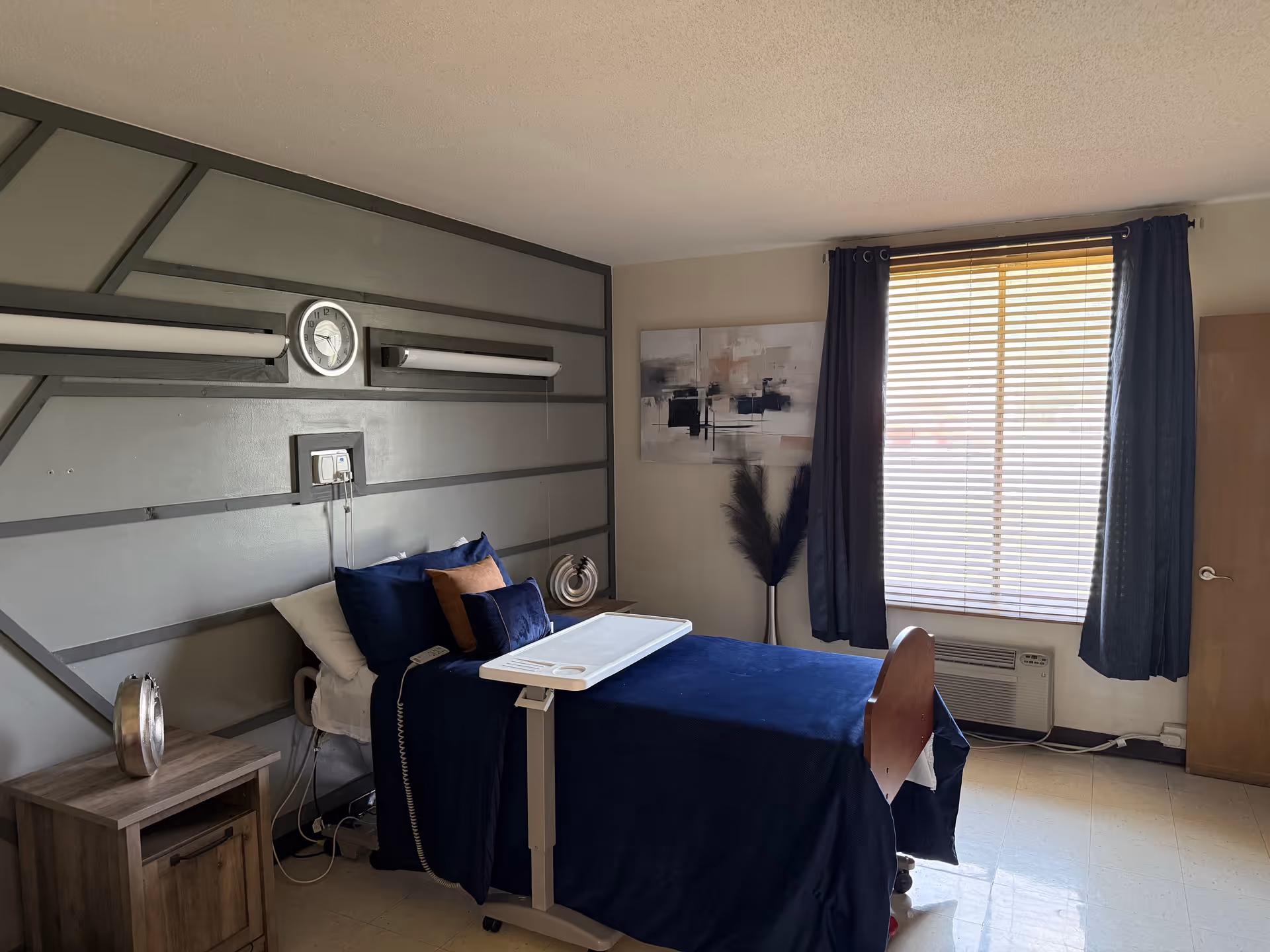 Hospital-style bedroom with adjustable bed, navy bedding, nightstand, wall decor, window with blinds.