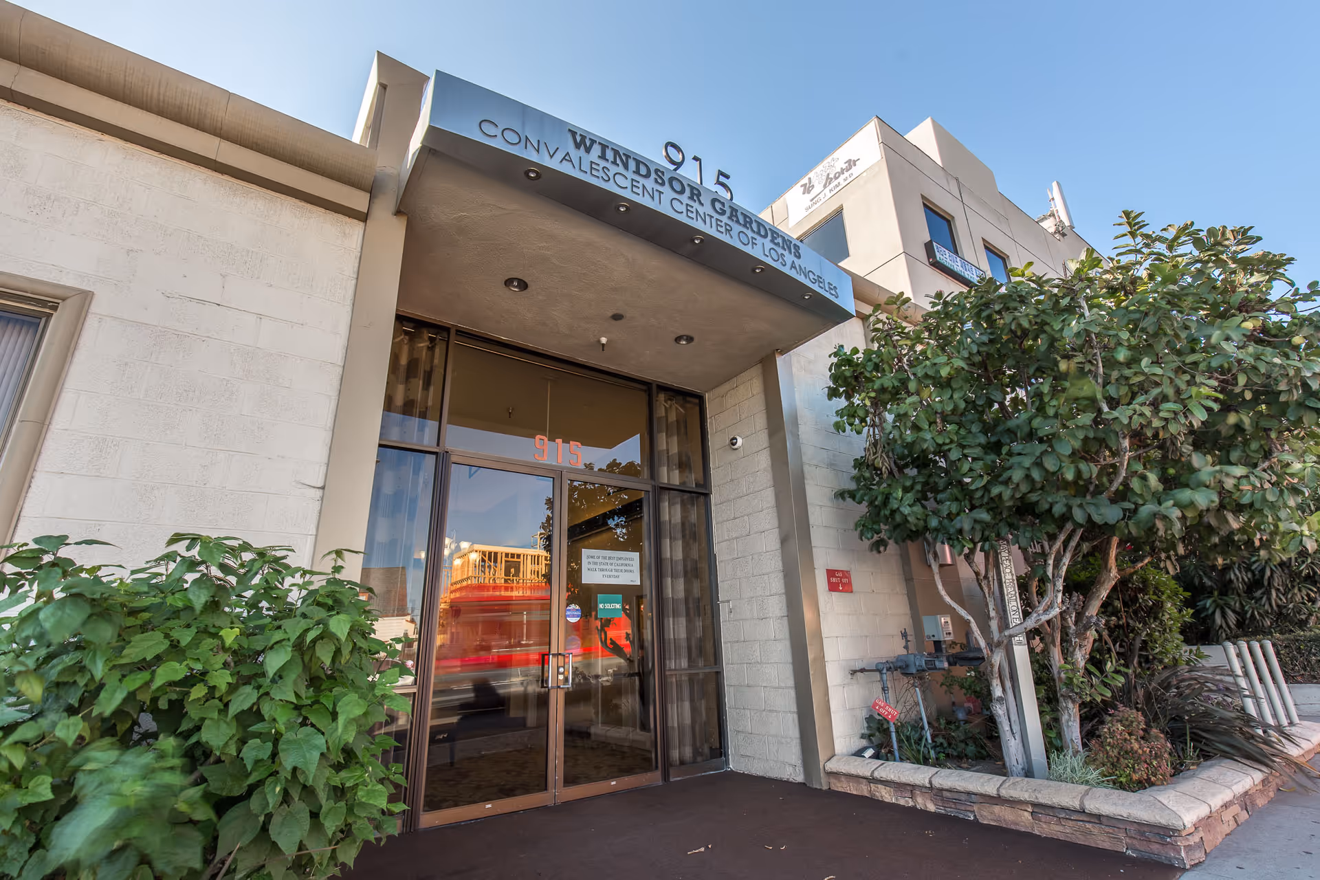Entrance of Windsor Gardens Convalescent Center of Los Angeles with glass double doors, a metal awning displaying the facility name and address number 915, surrounded by plants and trees.