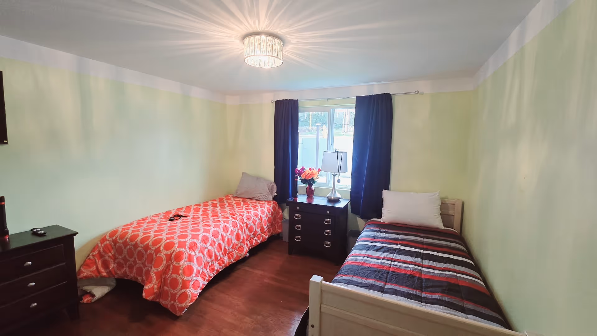 Two single beds with a dresser and window with blue curtains in a green-walled bedroom.