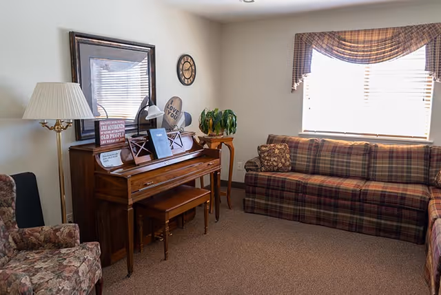A cozy living room with a wooden piano against the wall, a cushioned bench, a floral armchair, and a plaid sofa under a window with blinds and a valance. There is a floor lamp next to the piano, a small wooden side table with a plant, a wall clock, and decorative signs on the piano.