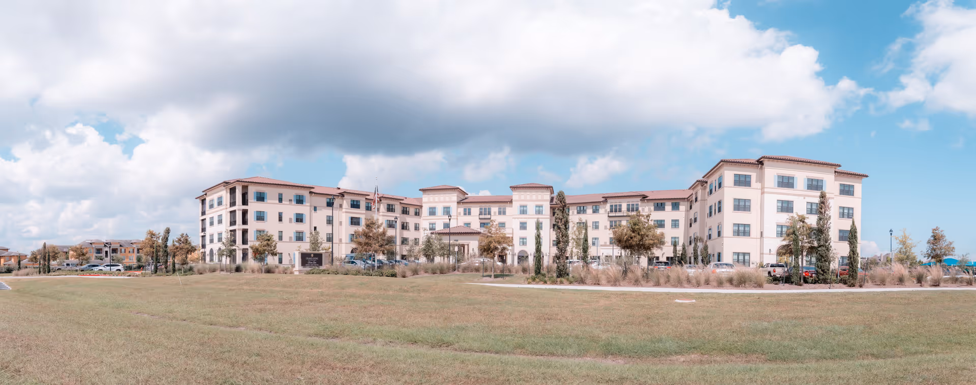 Front exterior of a large four-story Mediterranean-style senior living building set behind a grassy lawn under a partly cloudy sky.