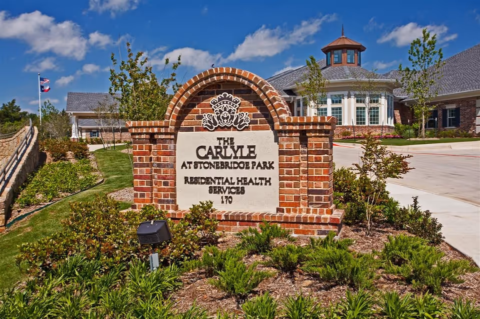 Brick entrance sign reading "The Carlyle at Stonebridge Park Residential Health Services" in front of the facility building under a blue sky.