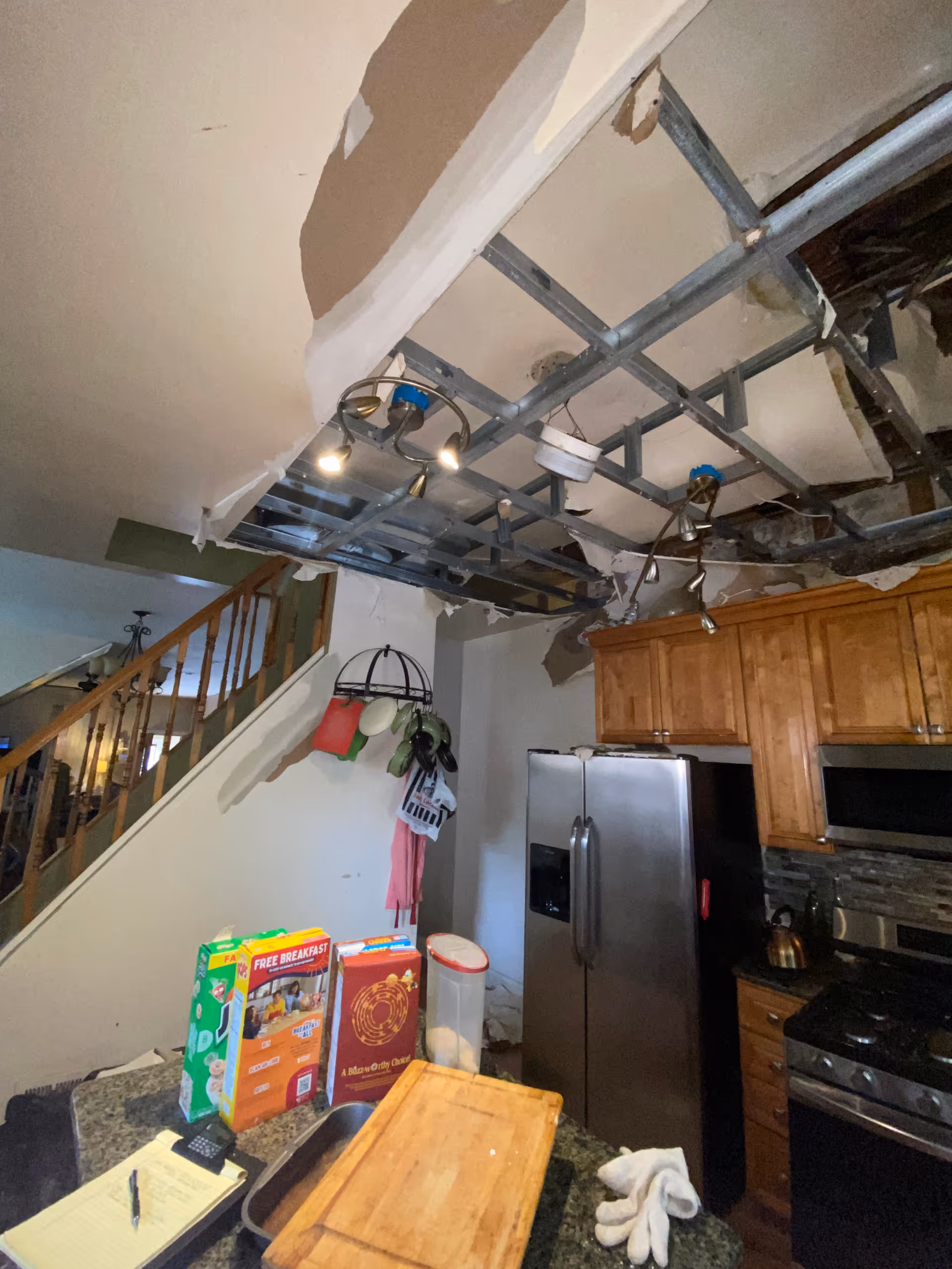Damaged kitchen ceiling with exposed metal framing above a fridge and wooden cabinets.