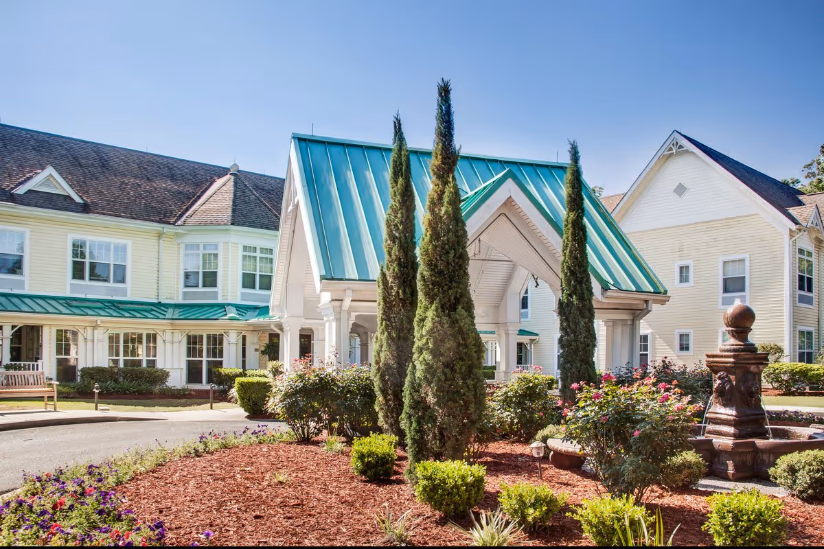 Front entrance of a senior living facility featuring a green metal-roofed porte-cochère, landscaped flower beds, and a decorative fountain.