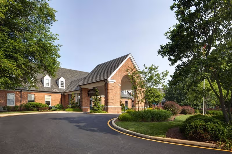 Brick senior living facility entrance with a covered porte-cochère, curved driveway, and landscaped trees and shrubs.