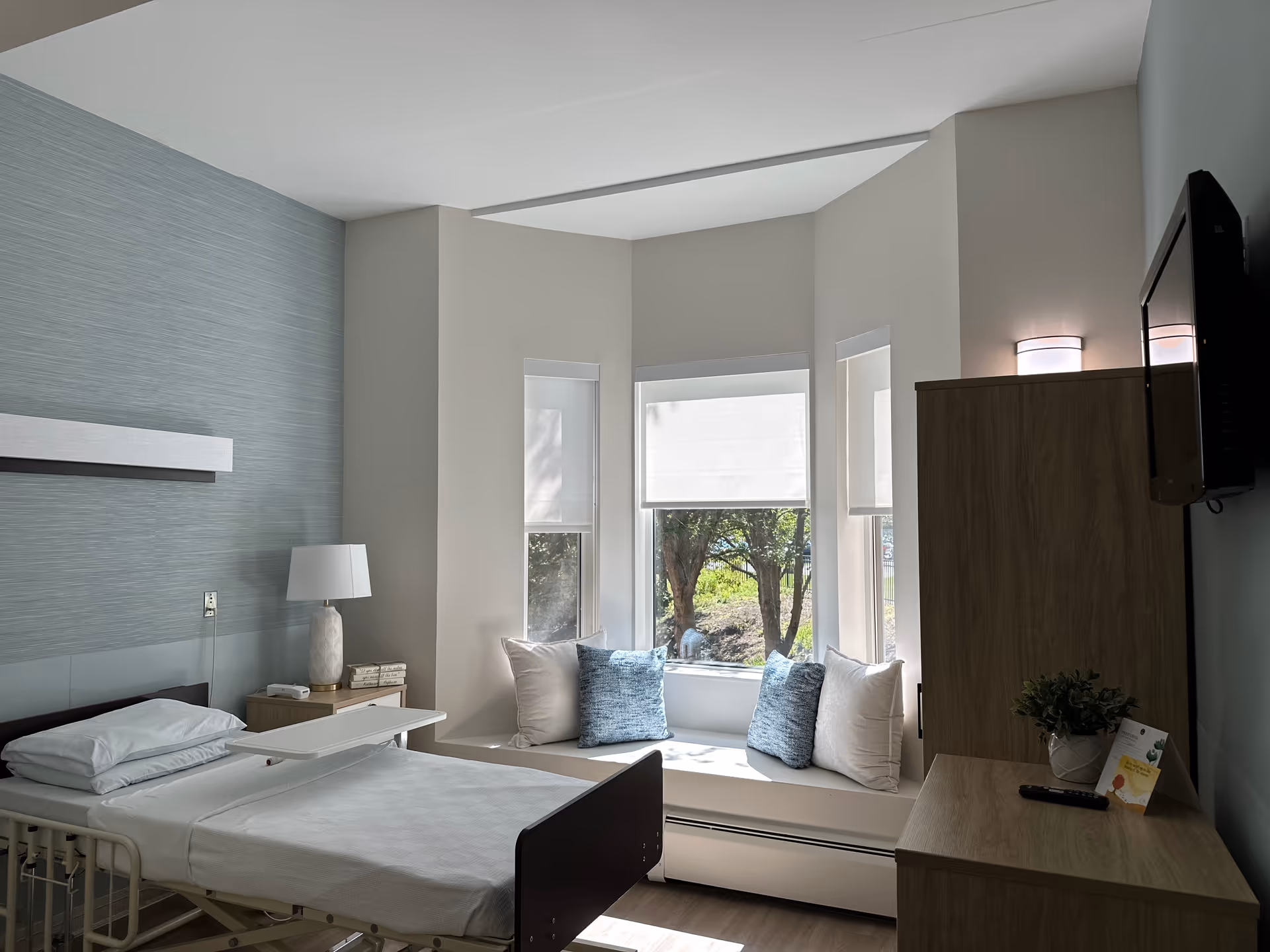 Bright patient room featuring an adjustable bed, window nook with cushions, and wood furniture.