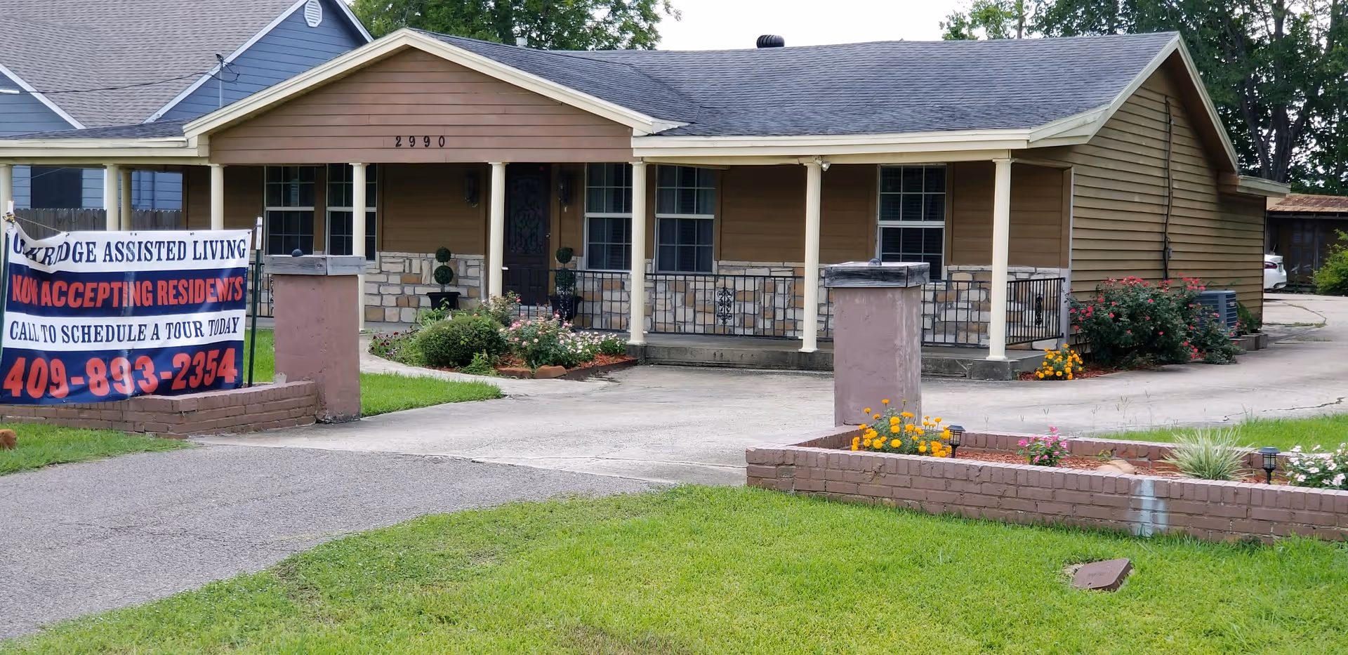 Front exterior of a single-story assisted living home with a covered porch, driveway, landscaped flower beds, and a sign advertising Oakridge Assisted Living accepting residents.