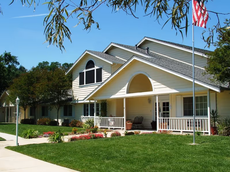 Front exterior of a light-colored assisted living building with a covered porch, manicured lawn, flowerbeds and an American flag.