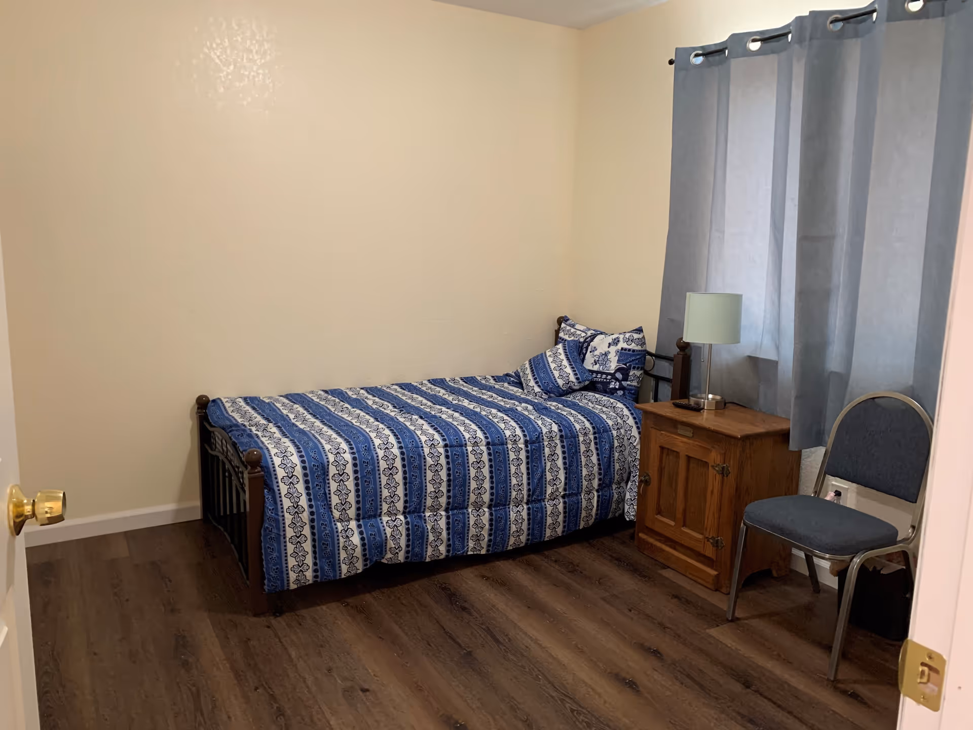 A small bedroom with a single bed covered in a blue and white patterned bedspread, a wooden nightstand with a lamp, a gray chair, and gray curtains covering the window. The floor is wooden and the walls are painted light beige.