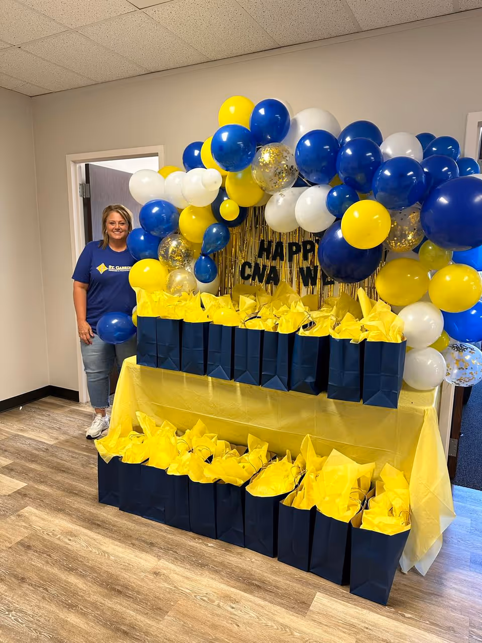 Staff member stands beside a blue and yellow balloon arch with gift bags and decor.
