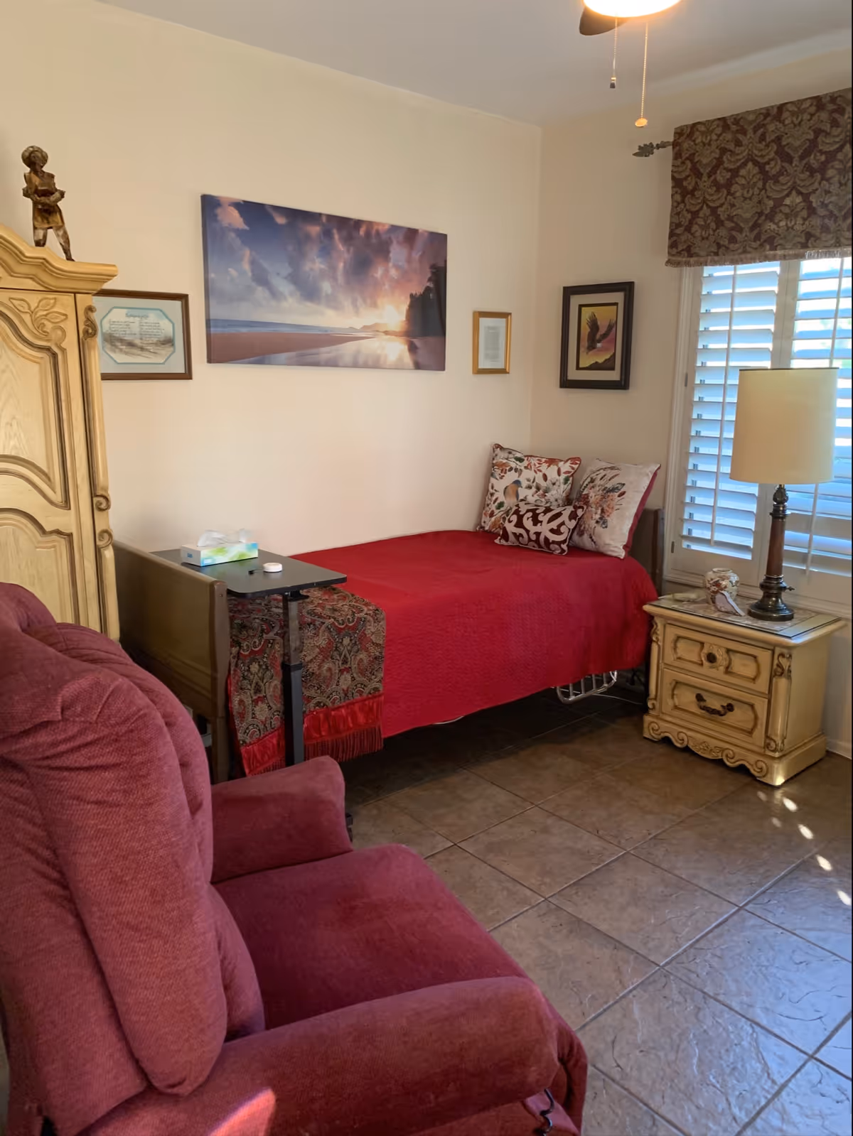 A furnished bedroom with red bedspread, armoire, nightstand, lamp, and upholstered seating.