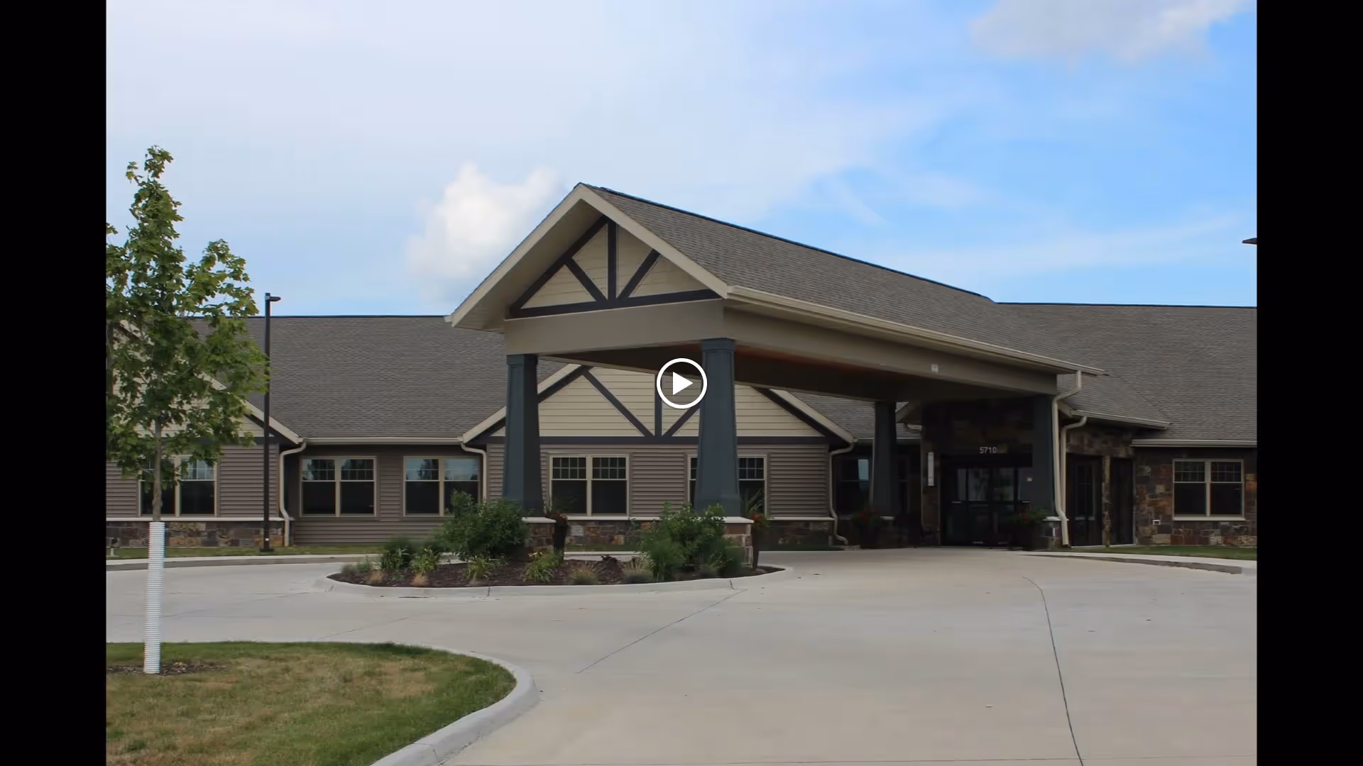 Front exterior view of a single-story senior living facility building with a covered entrance, beige and brown siding, stone accents, and a small landscaped area with shrubs and a young tree in front.