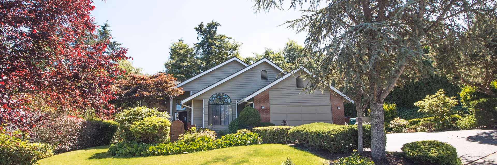 A single-story residential building with a gray exterior and a brick accent, surrounded by well-maintained landscaping including green bushes, trees, and a manicured lawn under a clear sky.