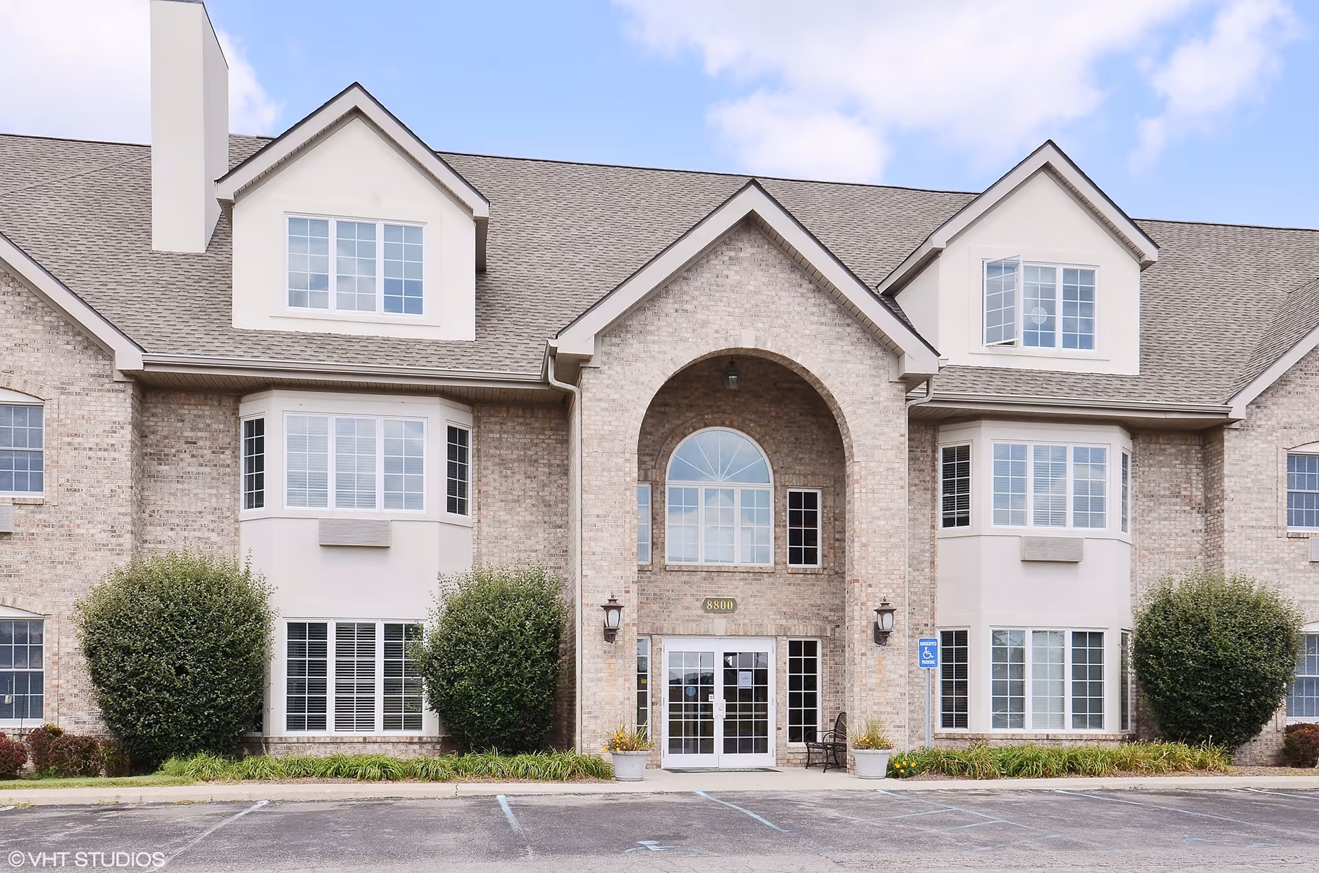 Front exterior view of a two-story senior living facility building with beige brick walls, multiple windows, and a central arched entrance with glass double doors. There are bushes and plants along the front, a parking lot with marked spaces, and a blue sky with some clouds above.