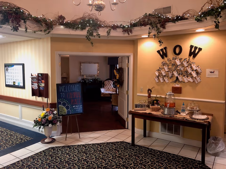 Interior view of a senior living facility entrance area with a decorative welcome sign on an easel, a table with refreshments including a beverage dispenser and hand sanitizer, a wall display with the letters 'WOW' and attached notes, floral arrangements, and a glimpse into a cozy sitting room with armchairs and a mirror.