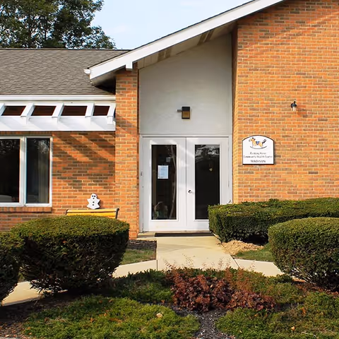 Brick building entrance with glass doors, manicured shrubs, and an exterior sign beside the doorway.