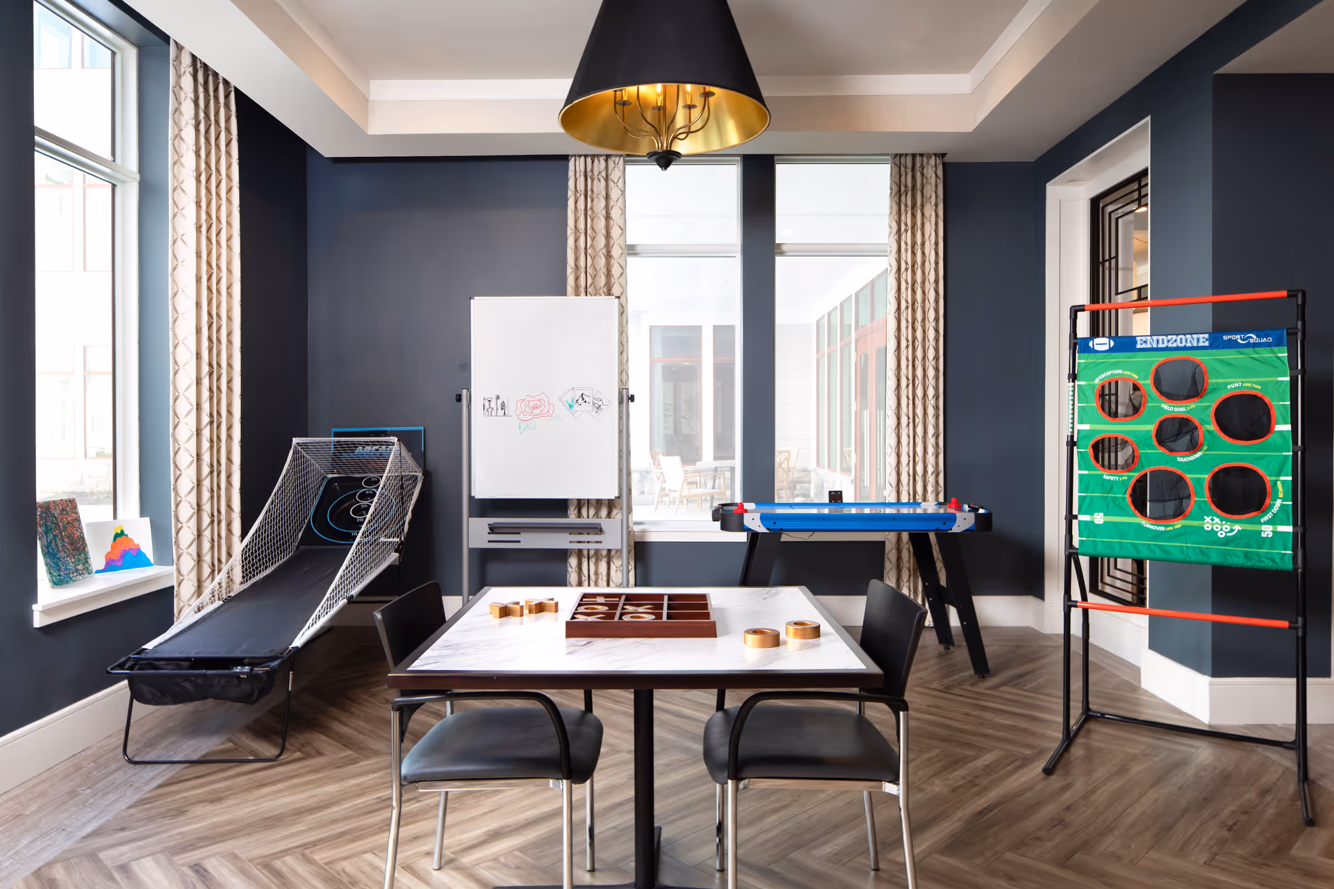 A bright activity room with dark blue walls and large windows with patterned curtains. The room features a table with two chairs and a tic-tac-toe game set on it. There is a basketball toss game on the left, an air hockey table in the back, and a green football toss game on the right. A whiteboard with drawings is positioned between the windows.