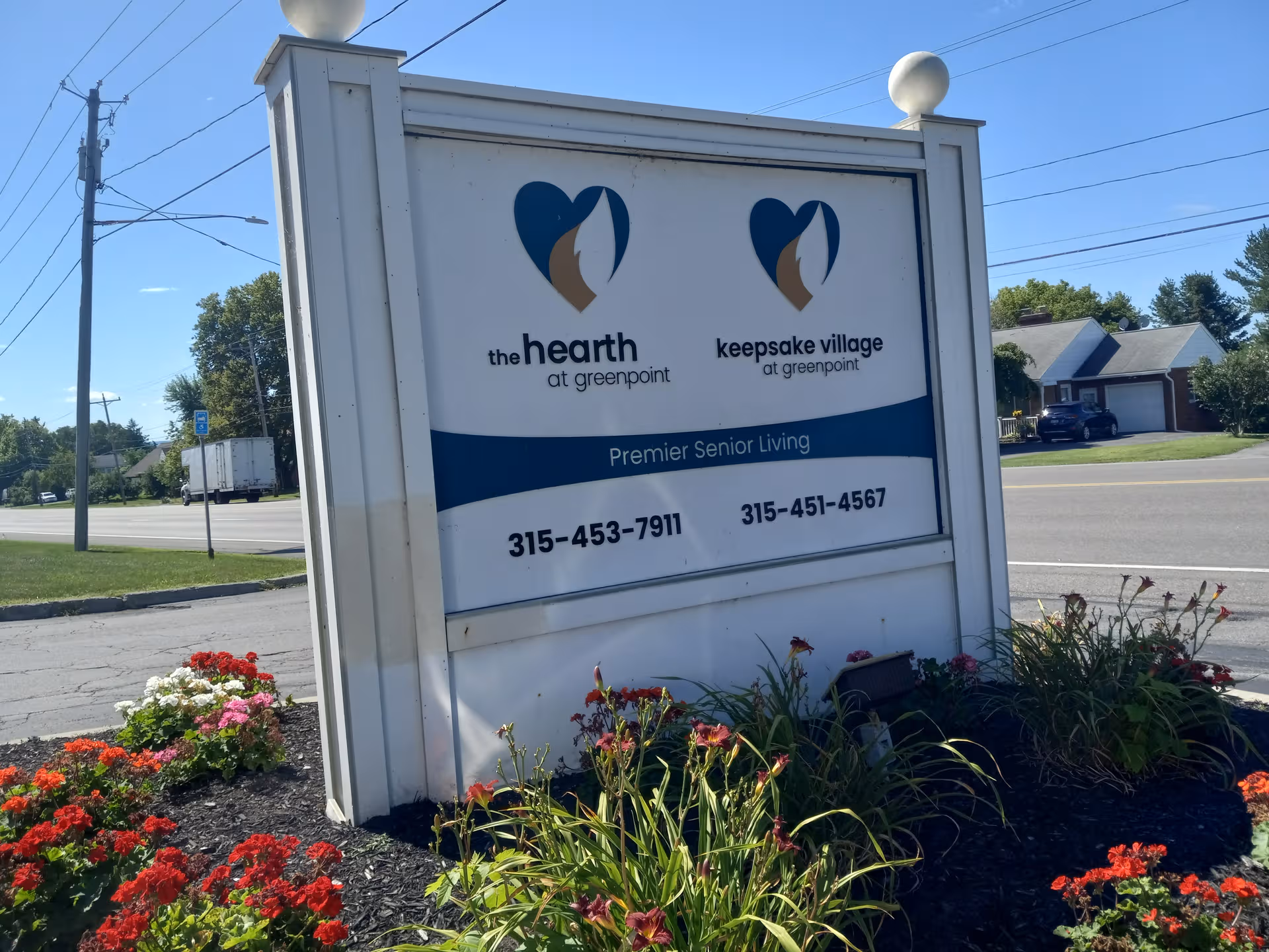 Outdoor sign for The Hearth at Greenpoint and Keepsake Village, with colorful flowers surrounding.