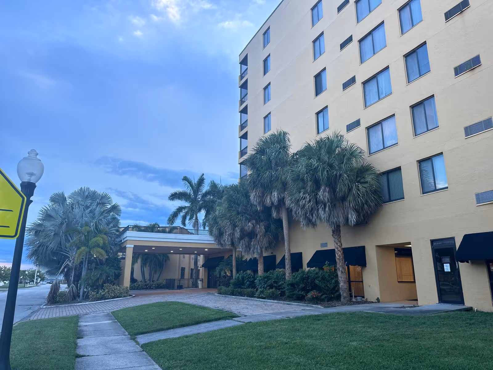 Exterior front view of a multi-story tan senior living building with a covered entrance, palm trees, and a sidewalk.