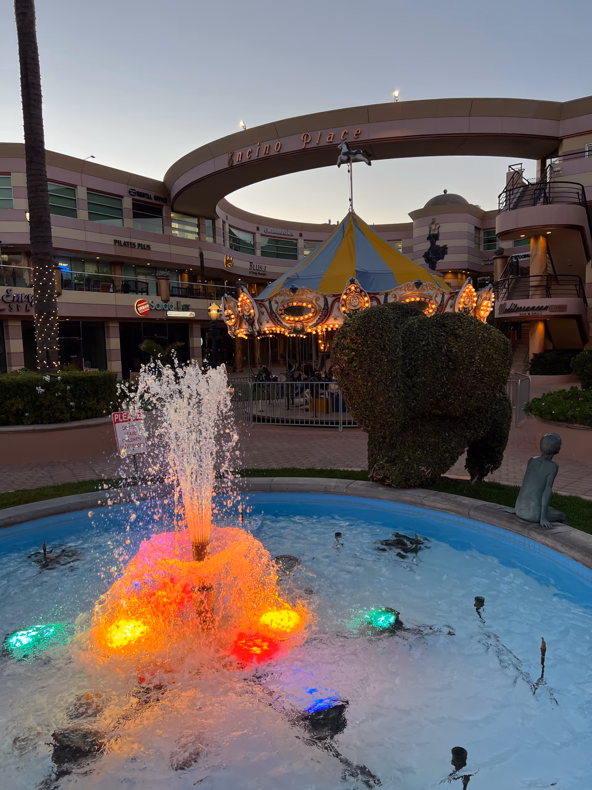 Outdoor courtyard featuring a lit fountain, carousel, and pastel building at dusk.