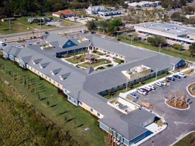 Aerial view of a large senior living facility building with a central courtyard featuring a gazebo and landscaped garden. The building has a grey roof and beige walls, surrounded by parking lots and green areas.