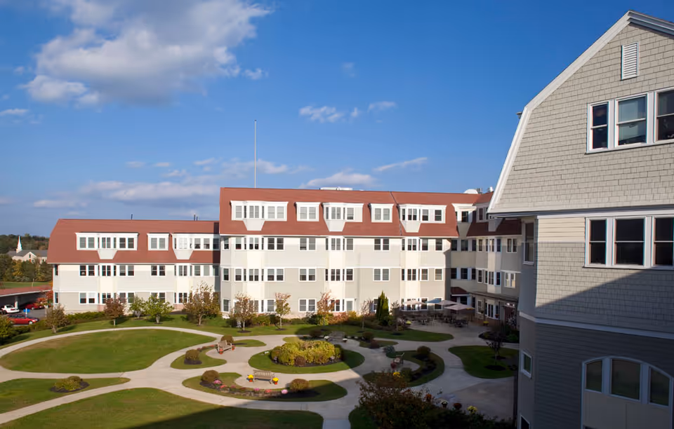Exterior view of a senior living facility named The Overlook, showing a multi-story building with red roofs and numerous windows. In front of the building is a landscaped courtyard with curved walkways, green lawns, benches, and small bushes under a partly cloudy blue sky.