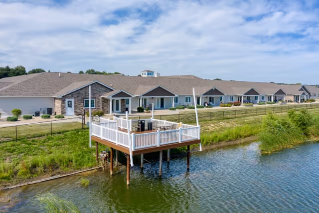 A senior living facility building with a long row of single-story units, situated next to a body of water. In the foreground, there is a raised wooden deck with white railings extending over the water, equipped with outdoor furniture. The sky is partly cloudy and the surrounding area is grassy with some shrubs.