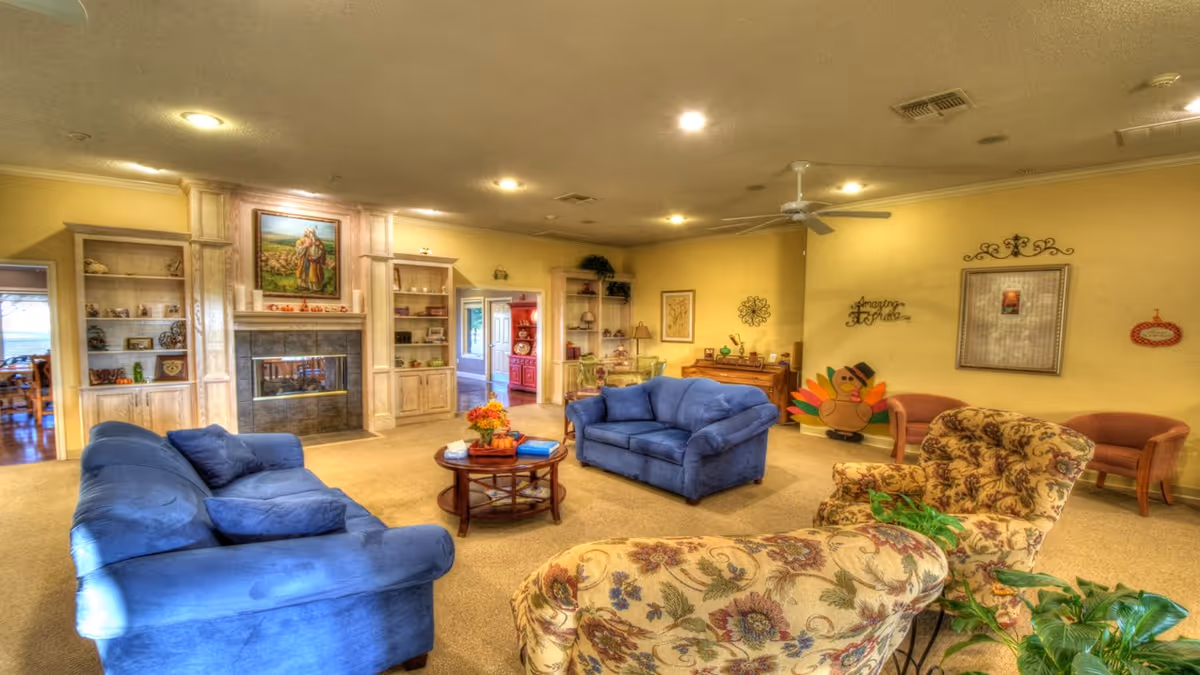 A spacious living room with two blue sofas, a floral patterned armchair, and a round wooden coffee table with a flower arrangement. The room features a fireplace with built-in shelves on either side, decorated with various items. The walls are painted yellow and adorned with framed artwork and decorative wall hangings. There is a ceiling fan and warm lighting creating a cozy atmosphere.