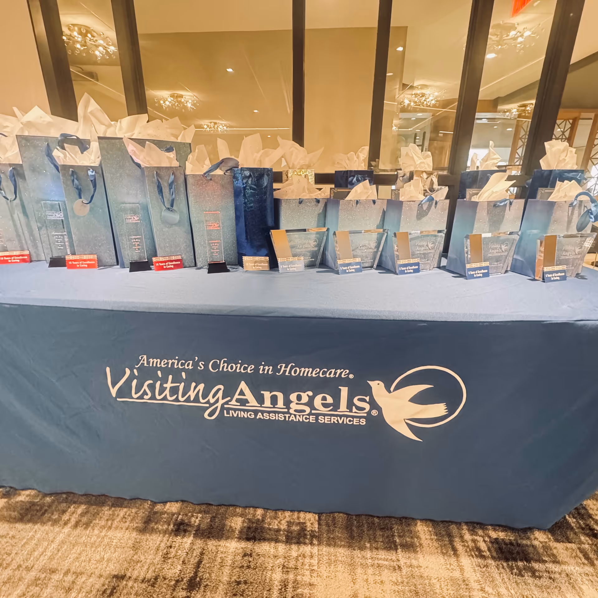 Table with blue tablecloth and gift bags bearing Visiting Angels logo indoors.
