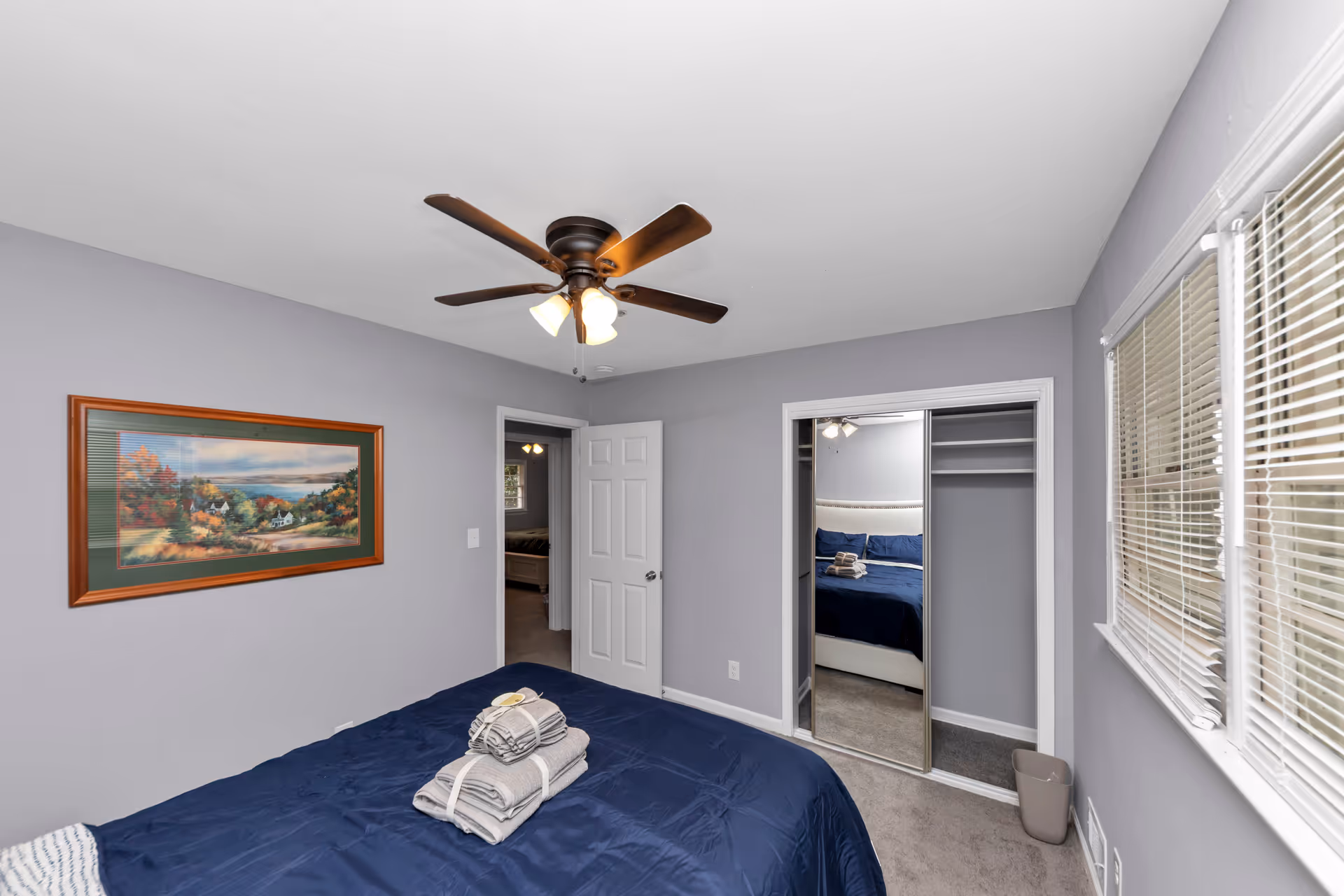 Bedroom with soft lavender-gray walls, blue bedding, ceiling fan, and mirrored closet doors.